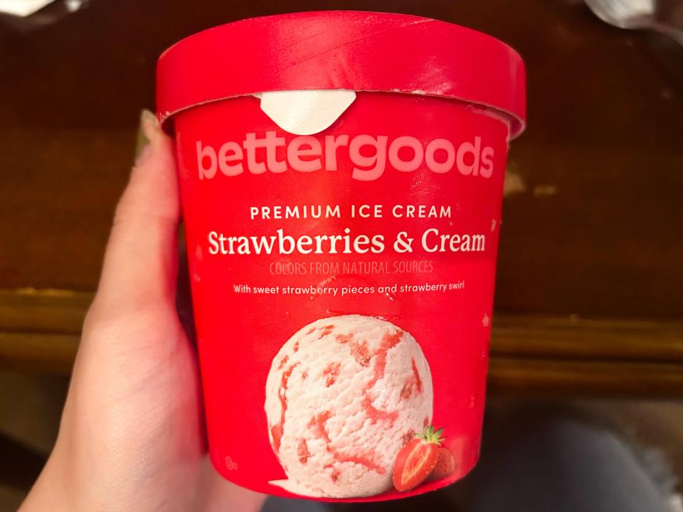 A hand holds a bright-red carton of ice cream with an image of strawberries and cream ice cream with red swirls in it on the container