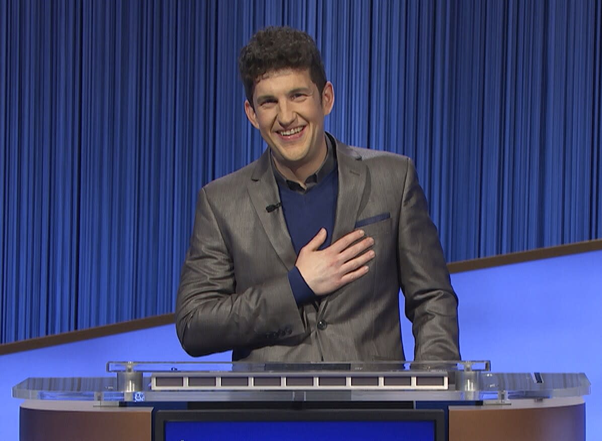 A man in a gray suit posing behind a podium with a hand on his chest