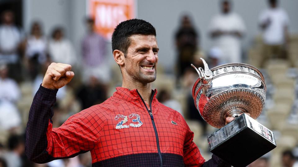 novak djokovic pumps his fist and holds the french open trophy, he wears a red and black jacket with the number 23 on the front