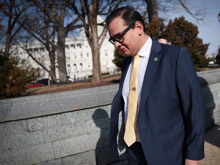 George Santos wearing black glasses, a blue suit and yellow tie while walking past reporters