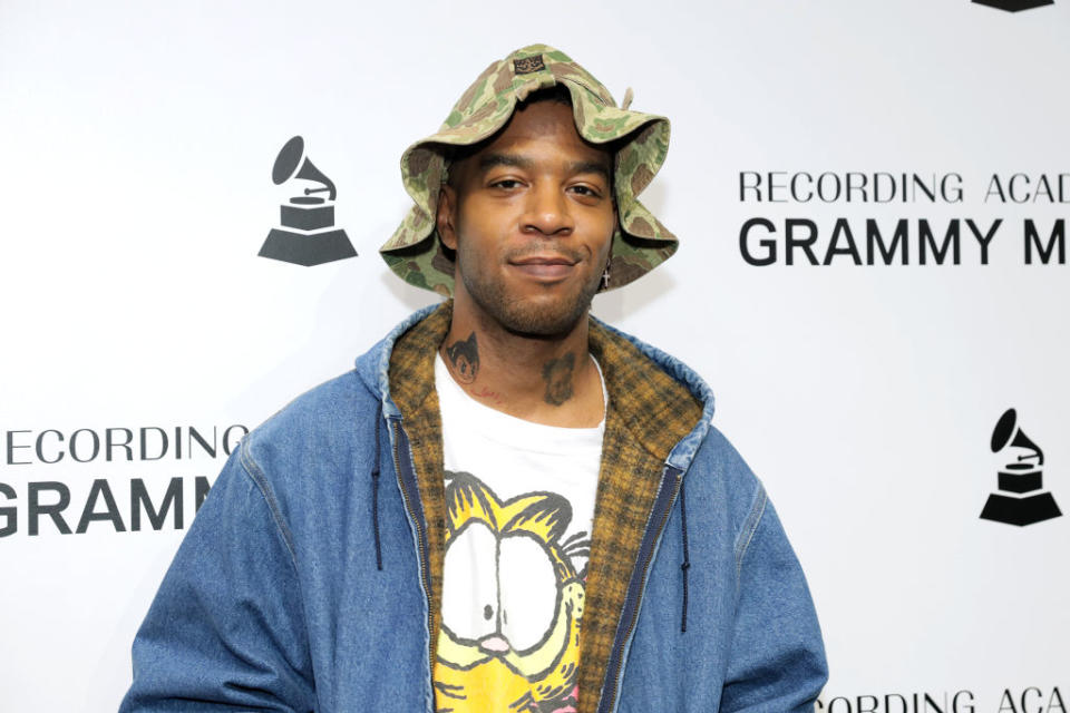 Closeup of Kid Cudi at a Grammy event wearing a denim coat, a t-shirt with Garfield on the front, and a camo print bucket hat
