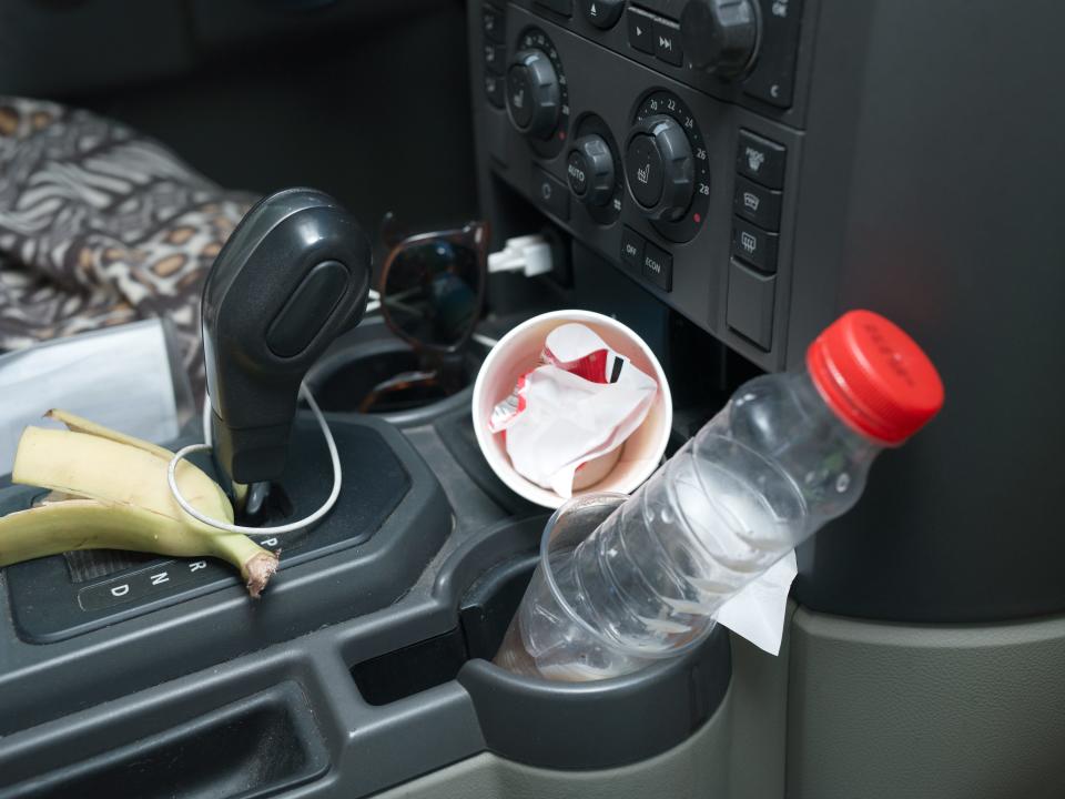 a banana peel, bottle, and cup filled with wrappers on the mid console of a car with a black interior