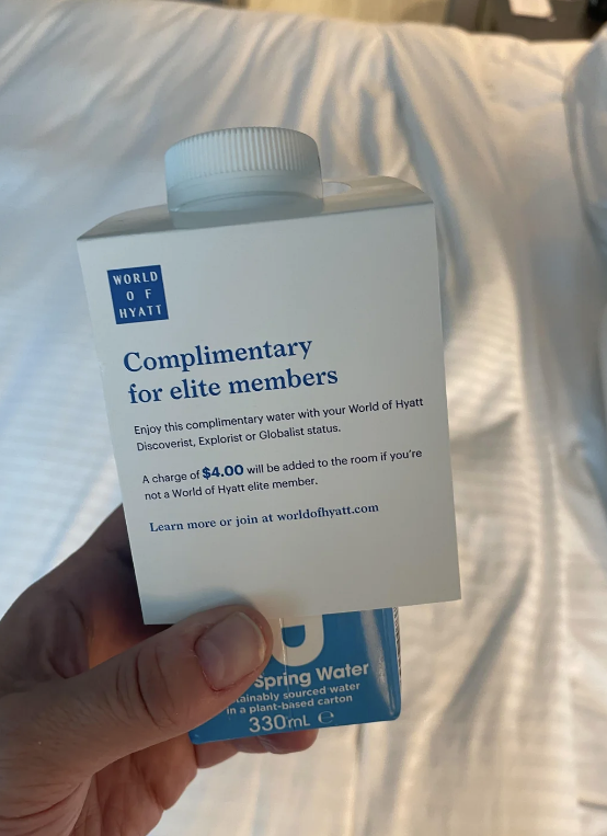 Hand holding a complimentary water carton by World of Hyatt, stating it's for elite members with Discoverist, Explorist, or Globalist status or for a charge of $4.00