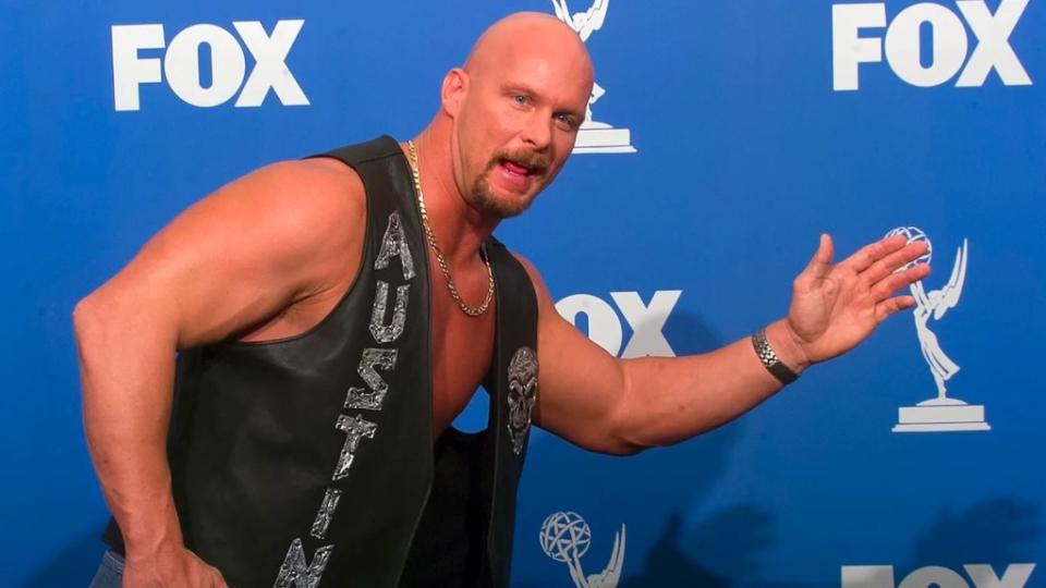 A bald man makes a fun running gesture in front of a backdrop with the Fox logo on it. He wears a vest that reads "AUSTIN."