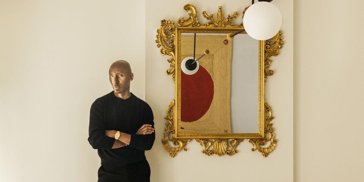 matthew harris, dressed in black, next to an antique gilded wall mirror, a low fireplace with logs, a modern wooden sculpted chair with a slender black metal back, and a light pendant with white and black orbs