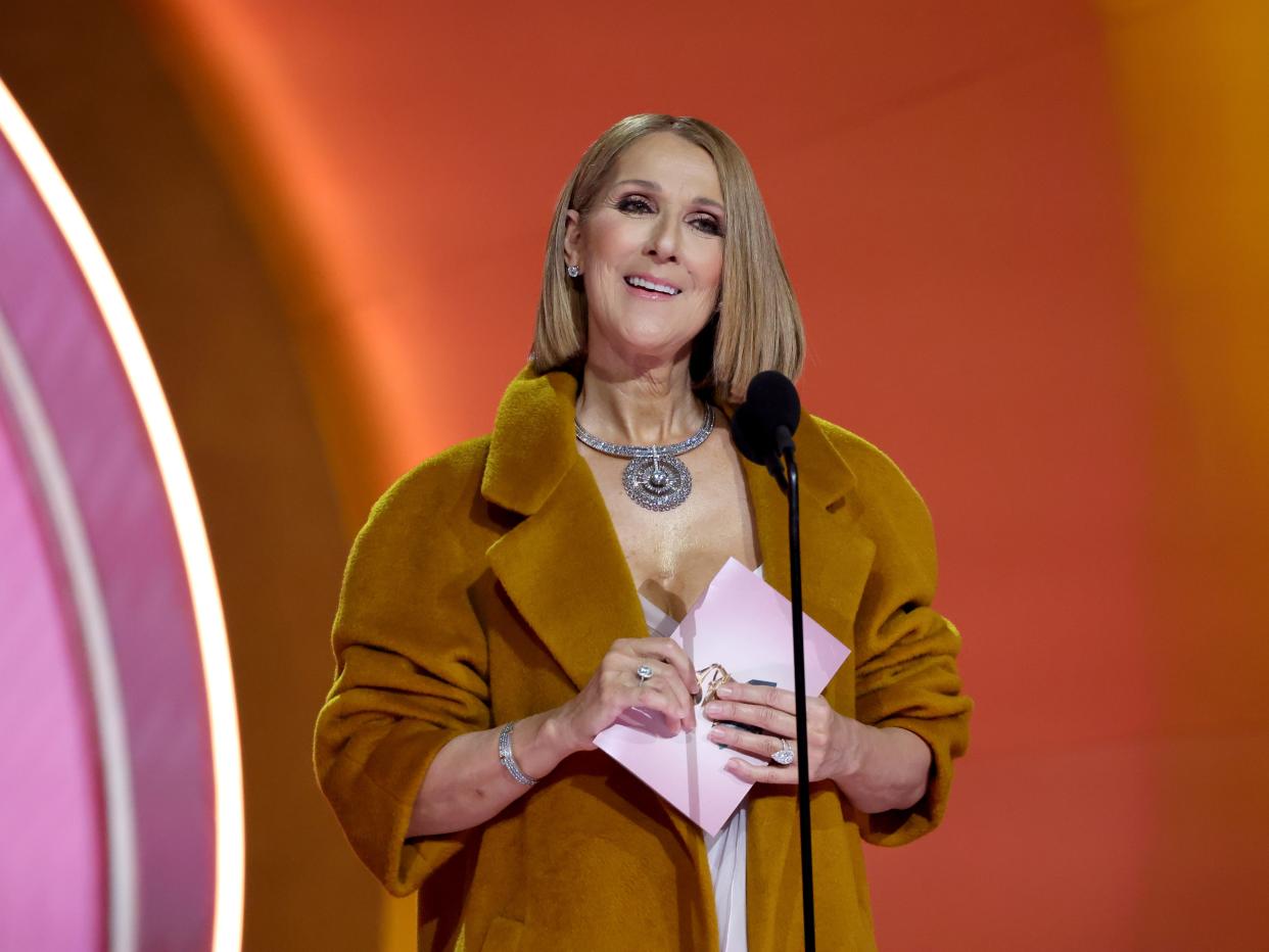 celine onstage at the grammy awards, standing in front of a microphone holding a pink envelope and wearing a brown overcoat