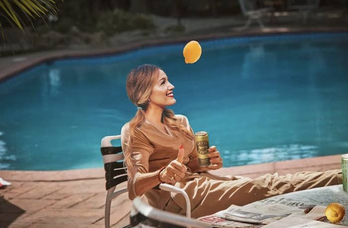 blake sitting by a pool with a lemon in the air, smiling, with a beverage can in hand. She's dressed in casual attire