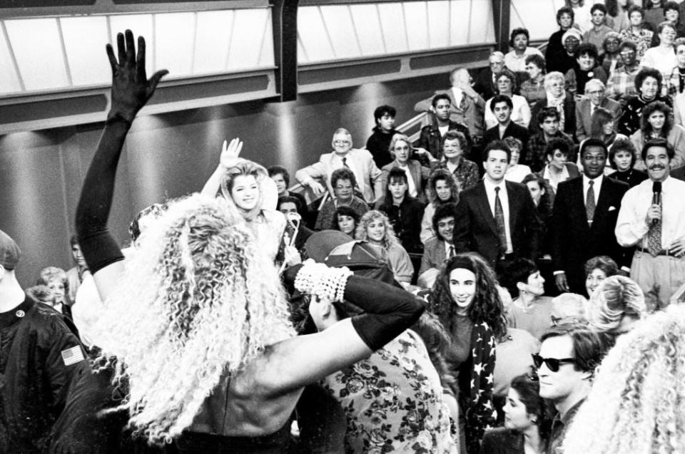 Crowd scene with an enthusiastic audience and performers, including a woman with curly hair and gloves waving to the crowd and another woman smiling nearby. Retro setting