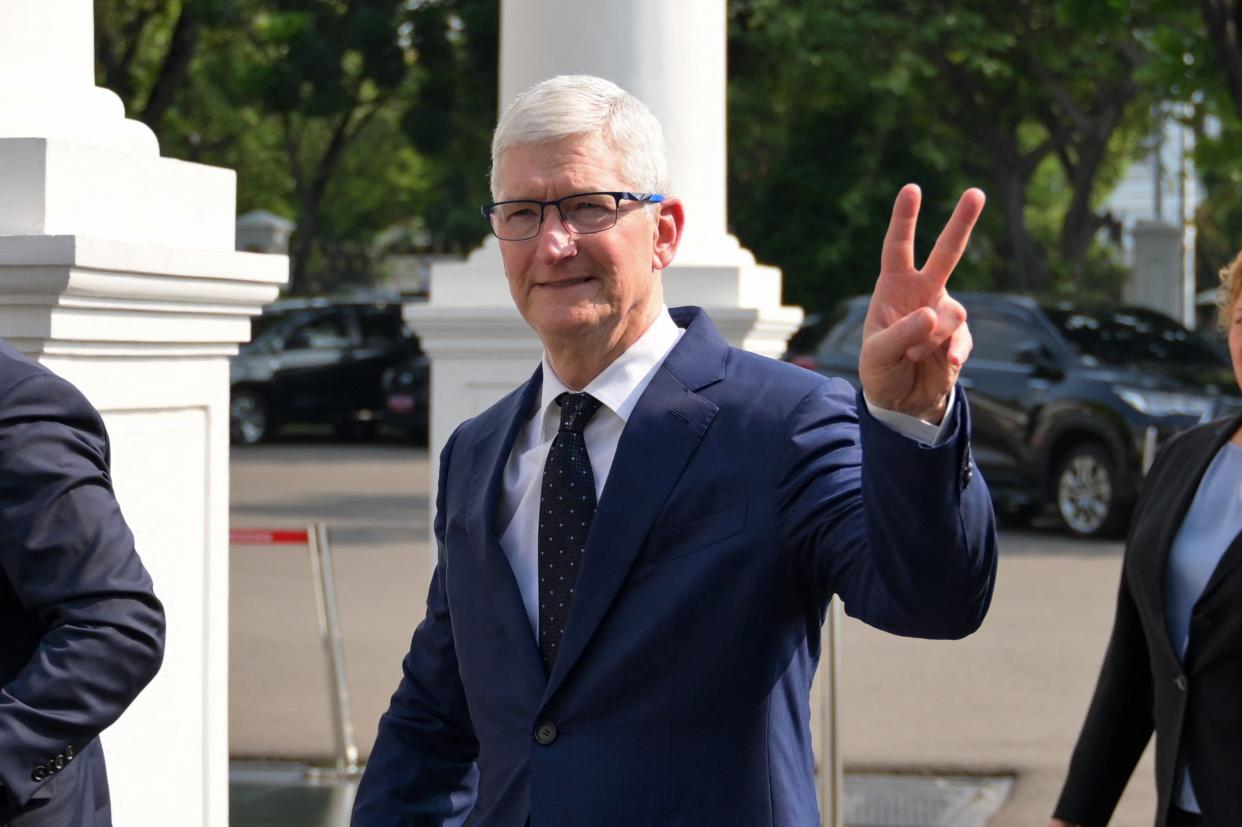 Apple CEO Tim Cook wearing a navy blue suit and making a peace sign with his hand whilst smiling