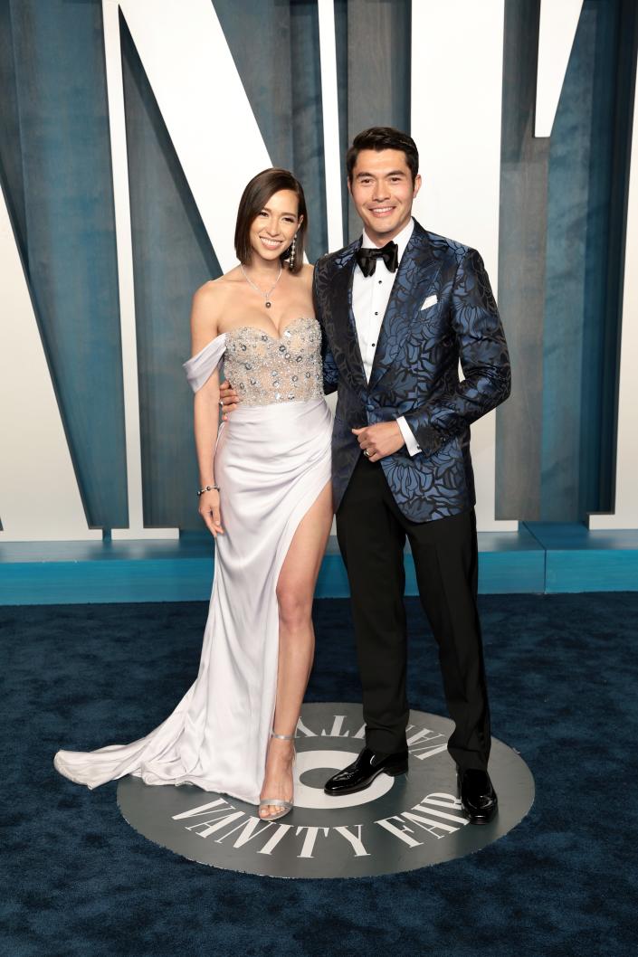 Liv in a light blue and silver dress.  The bodice is sheer and features large sequences and boning.  The off shoulder mini skeeves and thigh high slit skirt are made of satin.  Henry wears a dark blue shiny brocade blazer with a black bow tie and trousers.