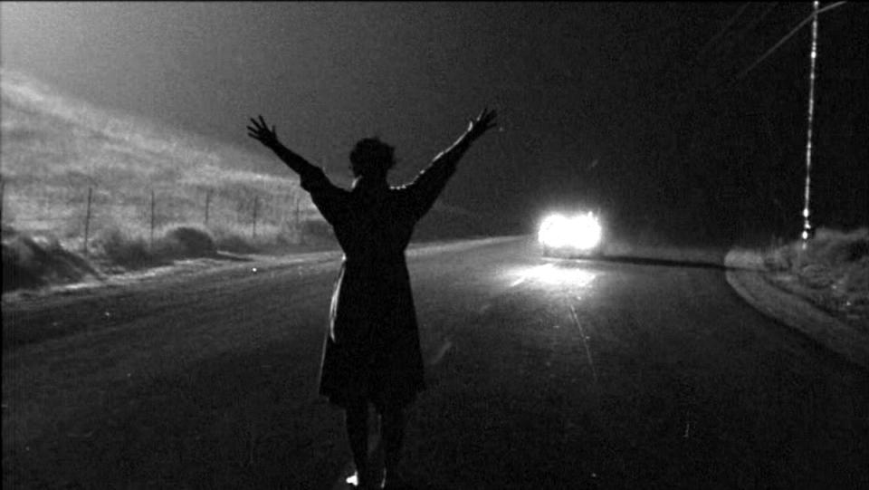 A woman stands in the middle of a street, holding her hands up to flag down an oncoming car in the film noir classic Kiss Me Deadly.