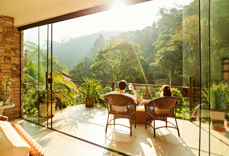 Two people sitting on wicker chairs on a patio, enjoying a scenic view of lush, sunlit greenery through large glass doors