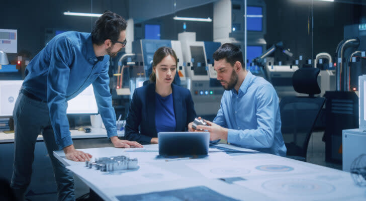 A photo of three people sitting around one end of a table, looking at a laptop screen.