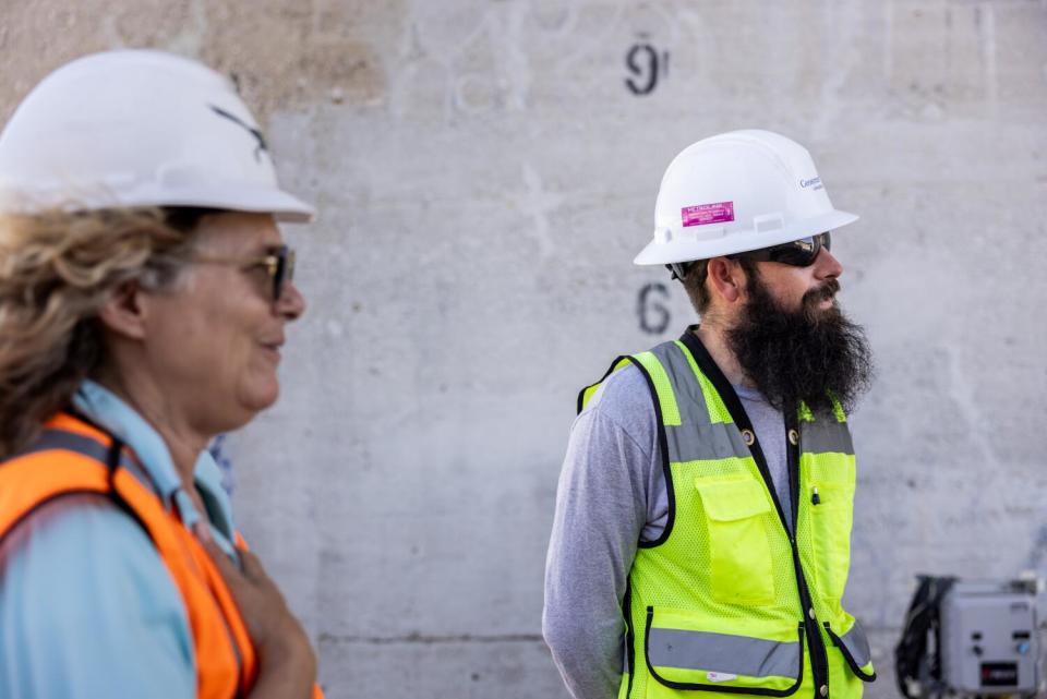 Lauren Bon, in orange safety vest and hard hat, stands next to Levi Walden, who has a big beard and sunglasses