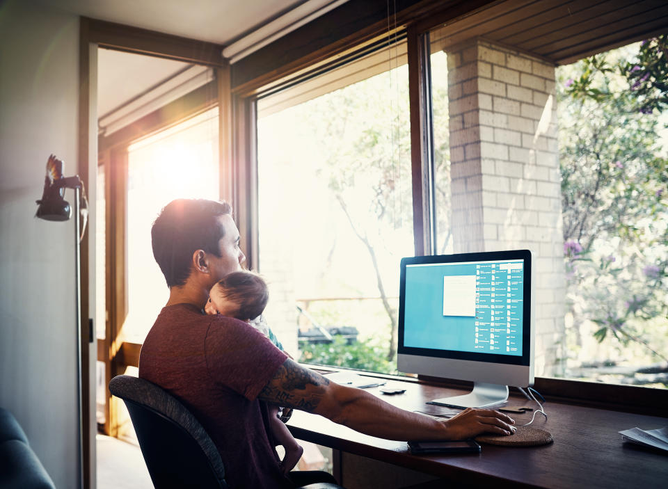 A young man working at home while holding his newborn baby son. (Source: Getty)