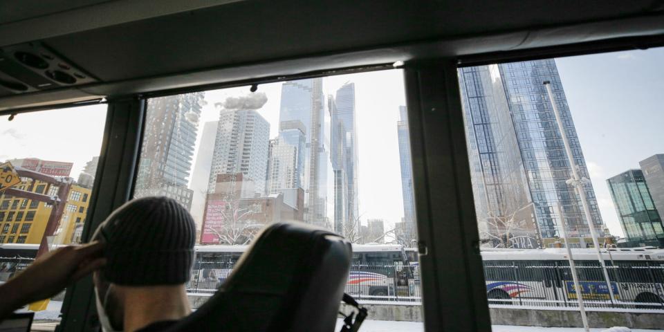 A view of tall buildings through bus windows. Someone wearing a beanie sits in the lower corner of the frame.