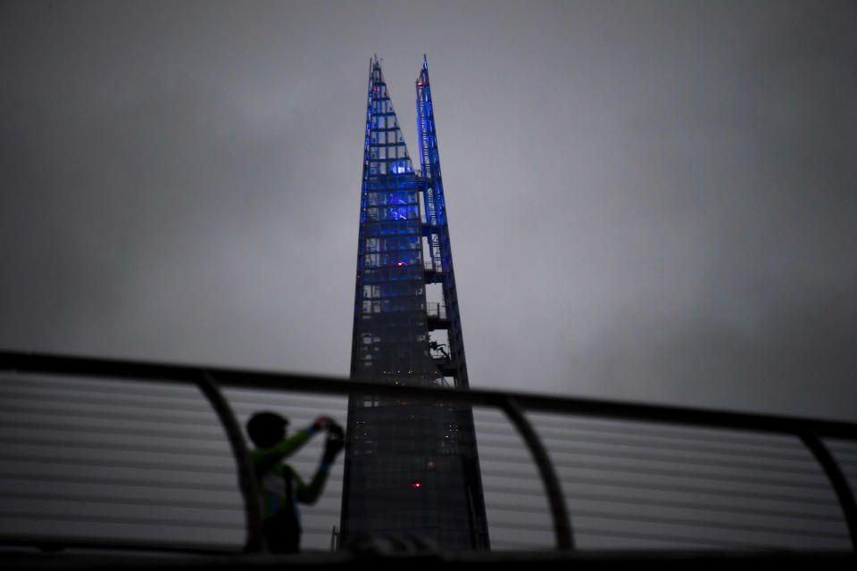 A man takes a picture on the Millennium Bridge as the Shard is lit up in blue in support of the National Health Service (NHS), as the capital is set to reopen after the lockdown due to the coronavirus outbreak, in London, Saturday, July 4, 2020. England is embarking on perhaps its biggest lockdown easing yet as pubs and restaurants have the right to reopen for the first time in more than three months. In addition to the reopening of much of the hospitality sector, couples can tie the knot once again, while many of those who have had enough of their lockdown hair can finally get a trim. (AP Photo/Alberto Pezzali)