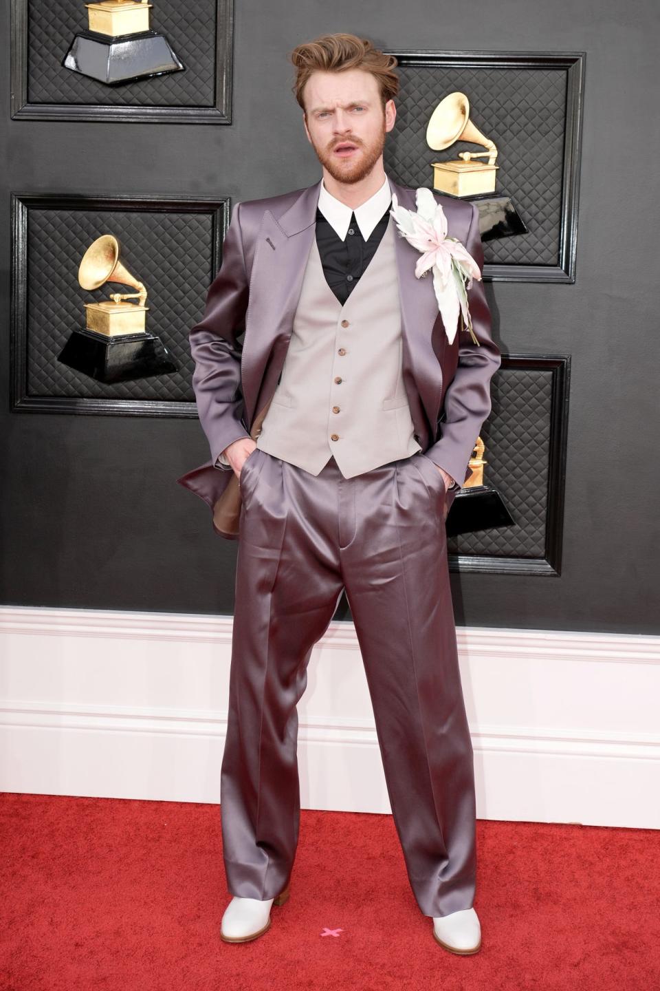 Finneas in a mauve and beige silk three piece suit with a black button down with a white collar under the vest. There's an oversized flower corsage attached to his jacket lapel. His dress shoes are cream-colored.