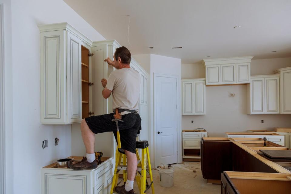 Man in work clothes installs white kitchen cabinets in new build kitchen. 