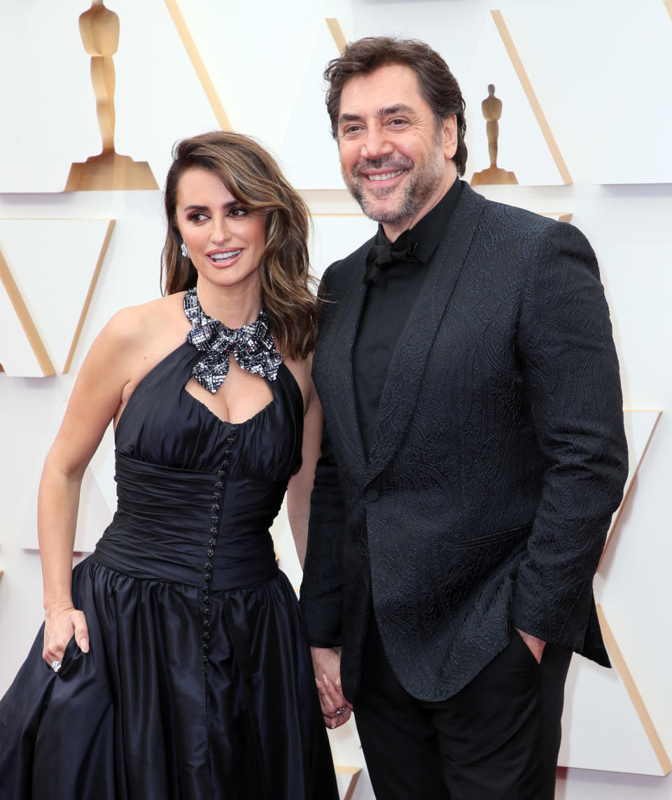 Two individuals pose together; one in a black halter-neck gown with an embellished neckline, the other in a black textured suit