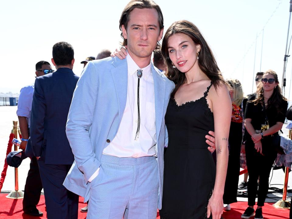 Lewis Pullman, in a powder blue suit, bolo tie, and brown boots, poses for photos with Rainey Qualley, in a black mini dress and heels, on the red carpet.