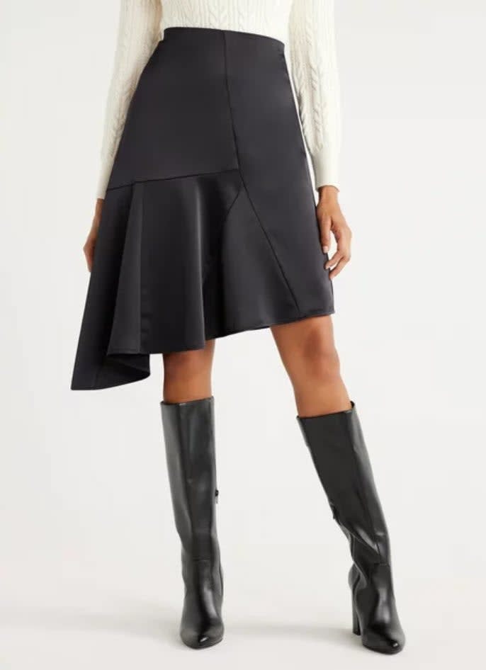Person in a white sweater, asymmetrical black skirt, and knee-high boots, cropped at the waist