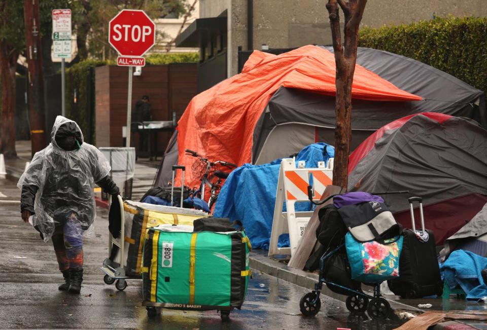 A woman in a plastic rain jacket holds onto a loaded trolley, with other packed belongings near tents