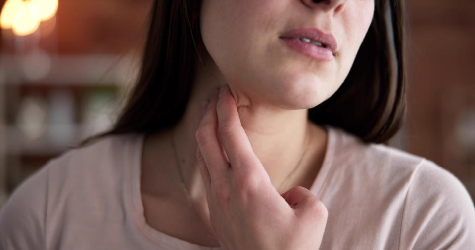 A woman touches her neck, appearing to check her pulse or feeling her skin. The image is closely focused on her face and hand