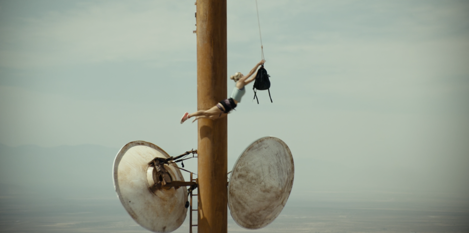 A blonde girl jumps from a satellite dish to a backpack hanging by a rope