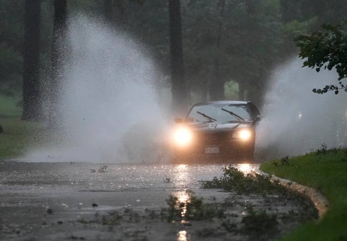 A car drives through a flooded road, splashing water as heavy rain continues. Trees and leaves are visible around the roadside