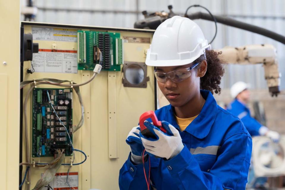 An electrician in a blue work suit and a white hard hat uses a tool to assess an electrical system.