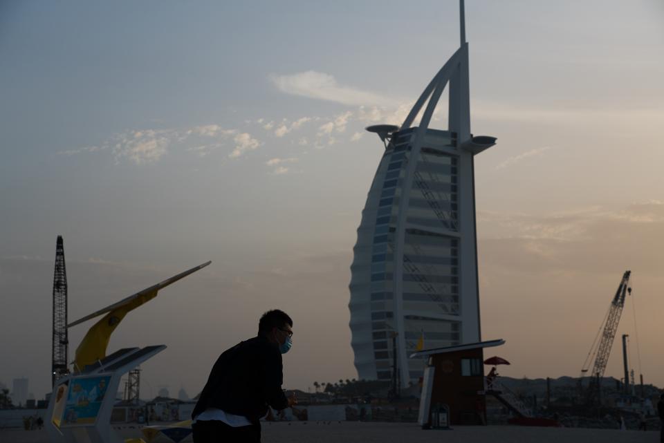A tourist wearing a surgical mask bends down to take a picture in front of the sail-shaped Burj Al Arab luxury hotel in Dubai, United Arab Emirates, Friday, March 20, 2020. The United Arab Emirates has closed its borders to foreigners, including those with residency visas, over the coronavirus outbreak, but has yet to shut down public beaches and other locations over the virus. (AP Photo/Jon Gambrell)