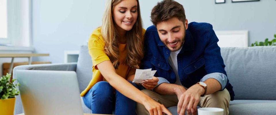 Happy young couple paying bills together and managing budget, sitting on the sofa and using calculator and laptop