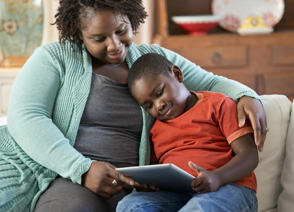 Woman and child sitting on a couch, both focused on a tablet the child is holding. The woman is wearing a cardigan, and the child is in a T-shirt