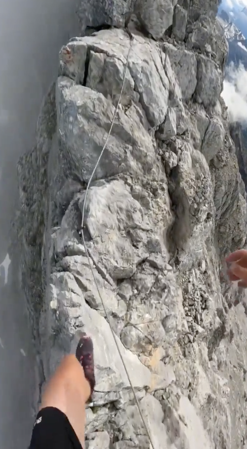 Person's legs visible walking on a narrow mountain ridge with a safety rope