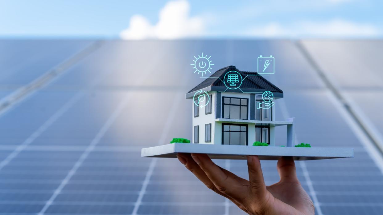  An architectural model of a house being lifted by a hand over huge solar panels, with icons representing energy concerns floating around it . 