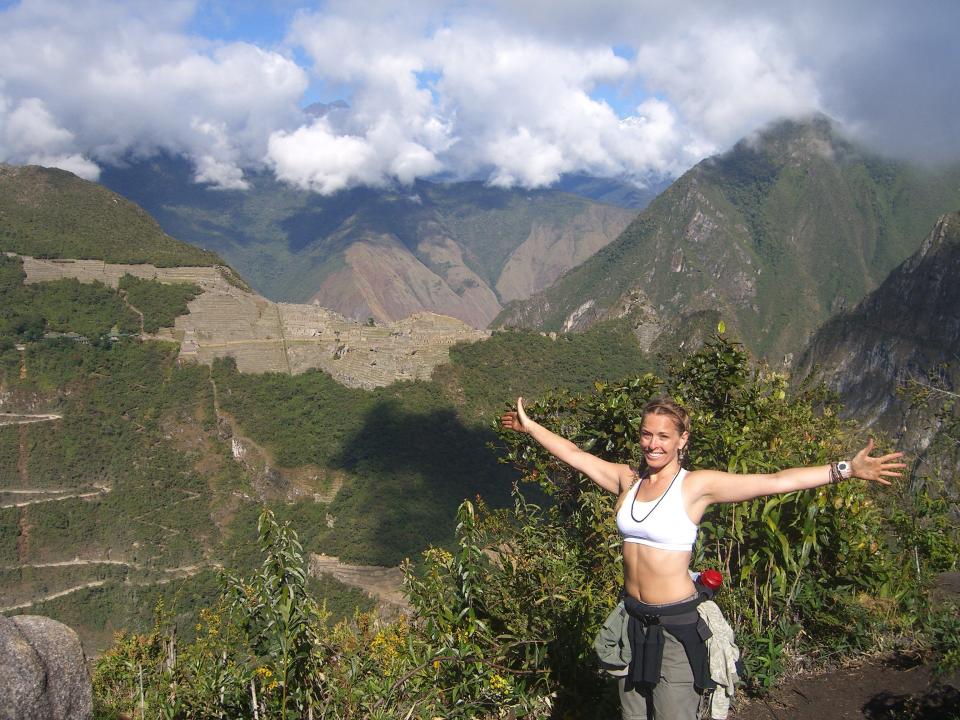 A woman stands with her arms out on a trail overlooking mountains on a cloudy day