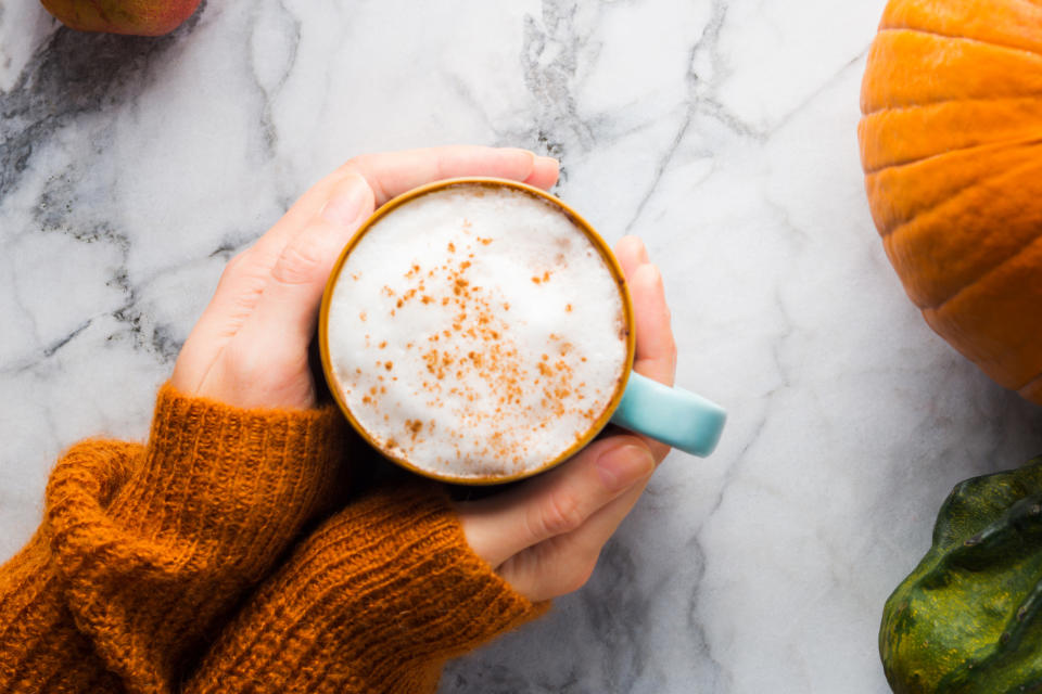 Hands holding a cup of coffee on a marble surface, surrounded by pumpkins