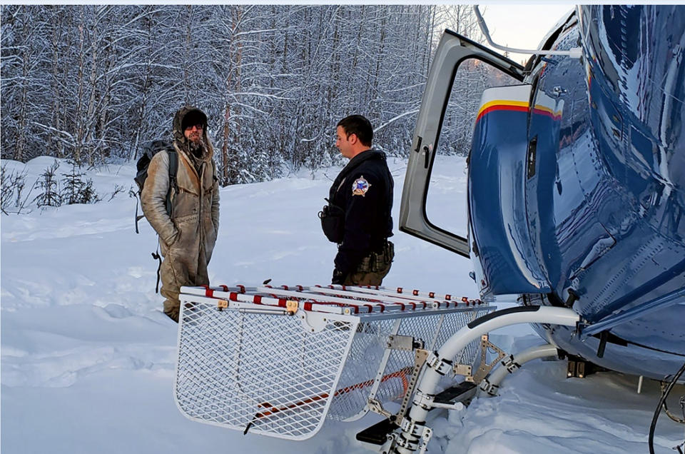 This Jan. 9, 2020 photo provided by Alaska State Troopers shows Tyson Steele, left, talking with Helo 3 Tactical Flight Officer Zac Johnson after Steele's rescue from outside Susitna Valley, Alaska. Troopers rescued Tyson Steele, 30, who survived in a makeshift shelter after his remote cabin burned last month, after spotting him and an SOS sign in the snow. (Alaska State Troopers via AP)