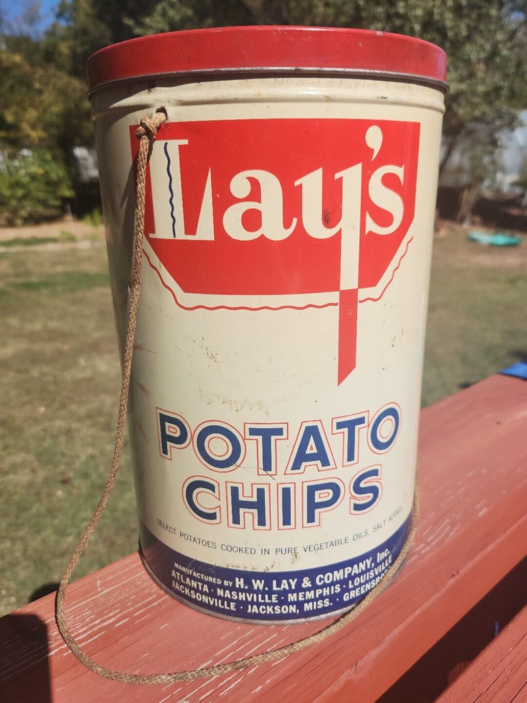 Vintage Lay's potato chips 1 lb. tin can with lid, white can with red and blue lettering and details, red lid, on a brick red fence with a lawn and trees blurred in the background