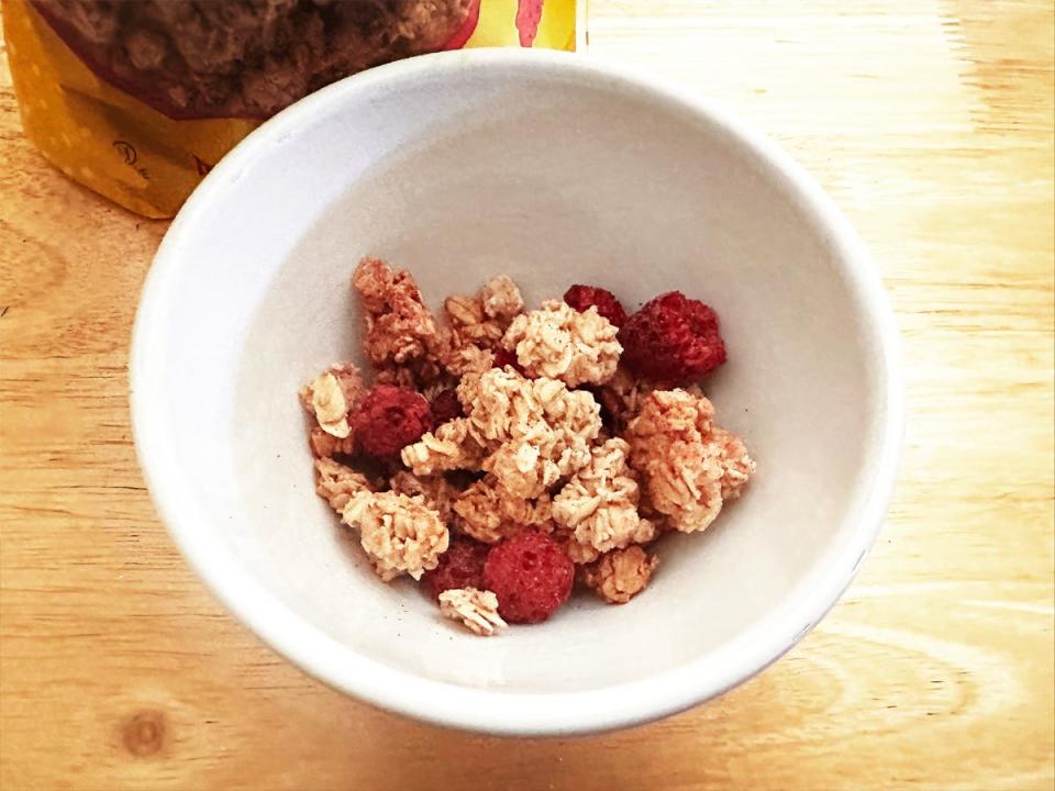 A white bowl filled with granola with pieces of raspberries in it on a wooden table