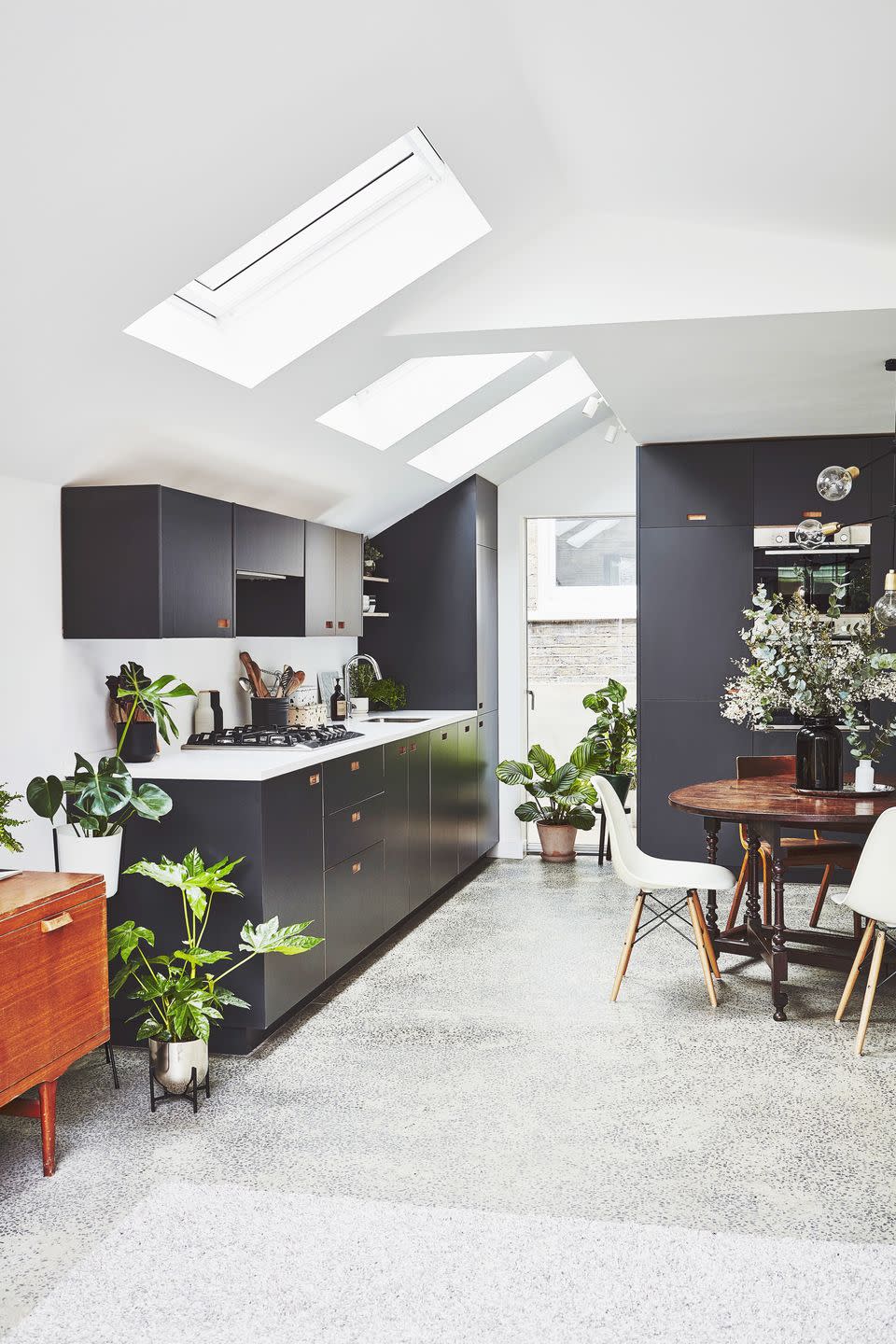 Gray kitchen units pulled from a bright white kitchen extension with terrazzo flooring, wooden dining table and potted plants.