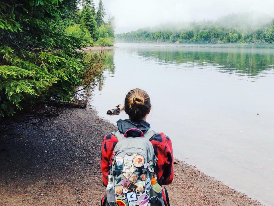Emily, wearing a flannel and a backpack covered in patches, sits on a log and looks out at foggy trees and water.