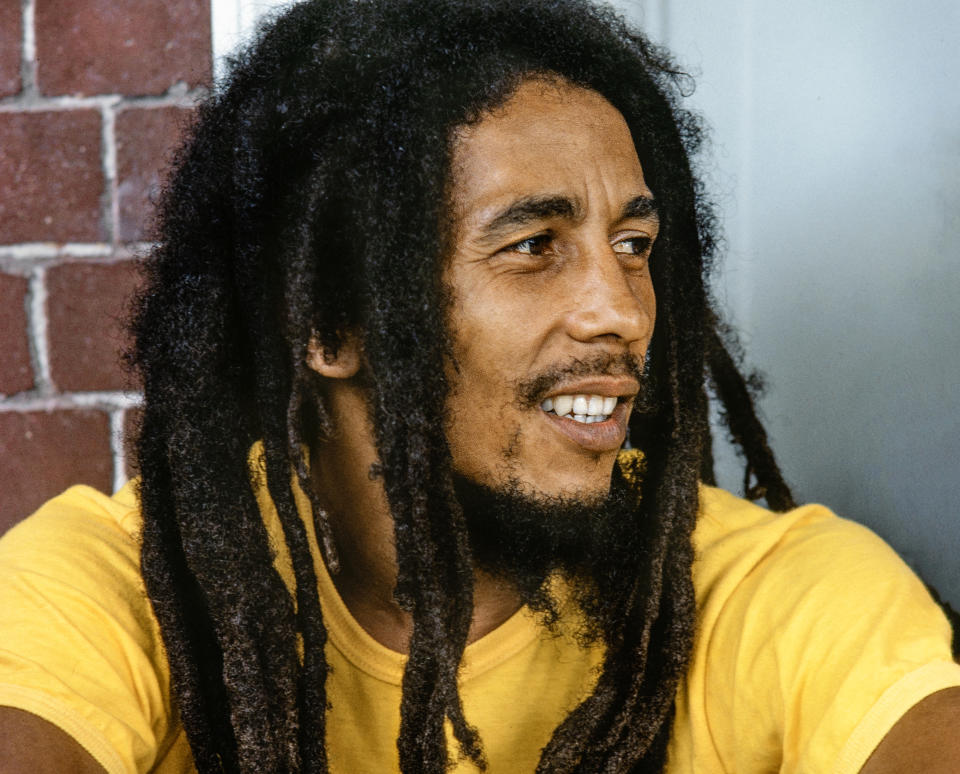 Bob Marley is sitting in a relaxed pose, wearing a simple shirt, and smiling to the right. His dreadlocks are prominently featured