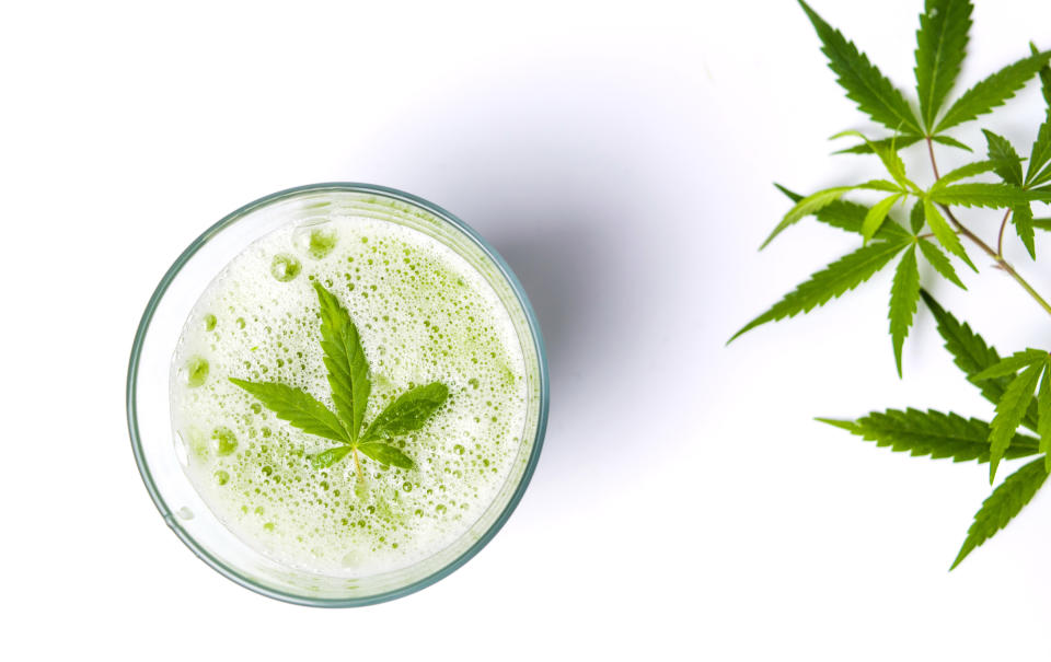A cannabis leaf sitting atop carbonation in a glass, with cannabis leaves to the right side of the glass.