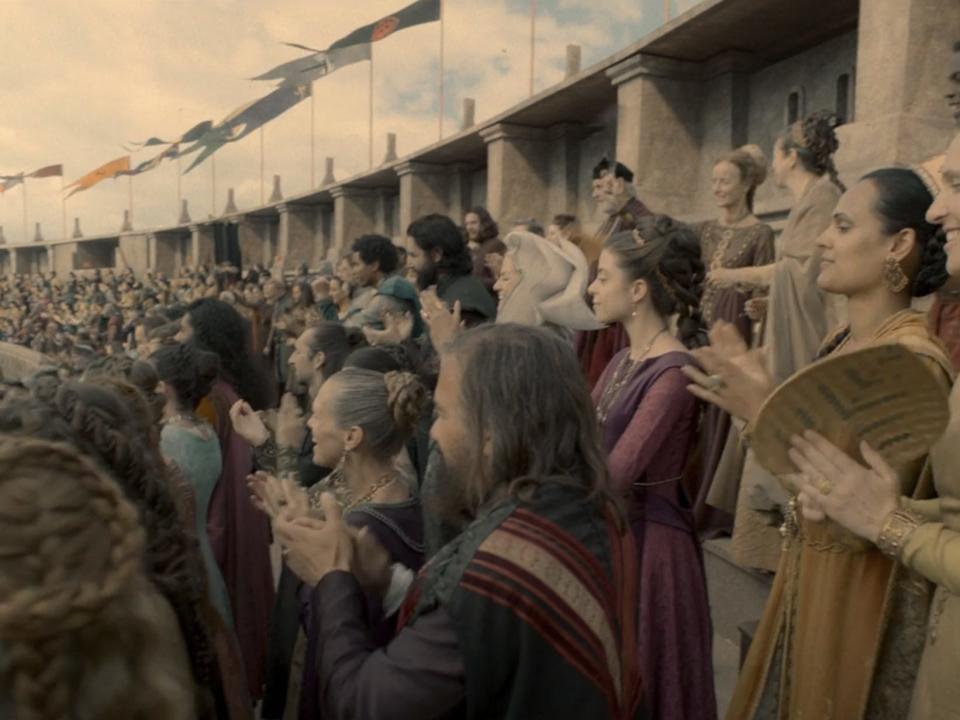 A crowd of spectators at a medieval tournament, with gold, pink, blue and other colored outfits and flags.