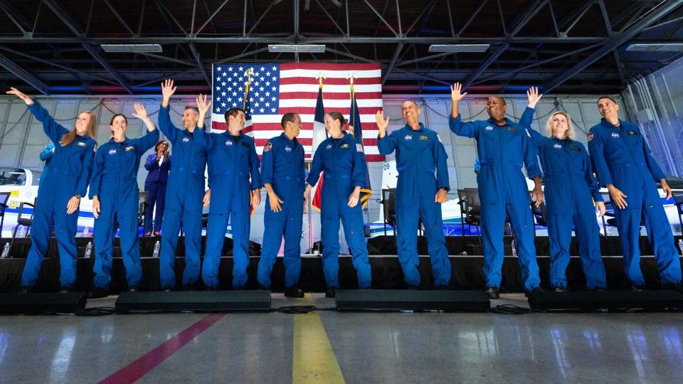 10 astronaut candidates stand in a row in a hangar, in front of an american flag and t-38 jets