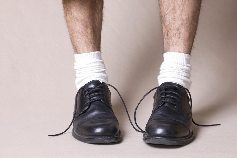 Close-up of a person's legs wearing white socks and black dress shoes with untied laces. The focus is on the shoes and socks, illustrating a casual or unfinished look