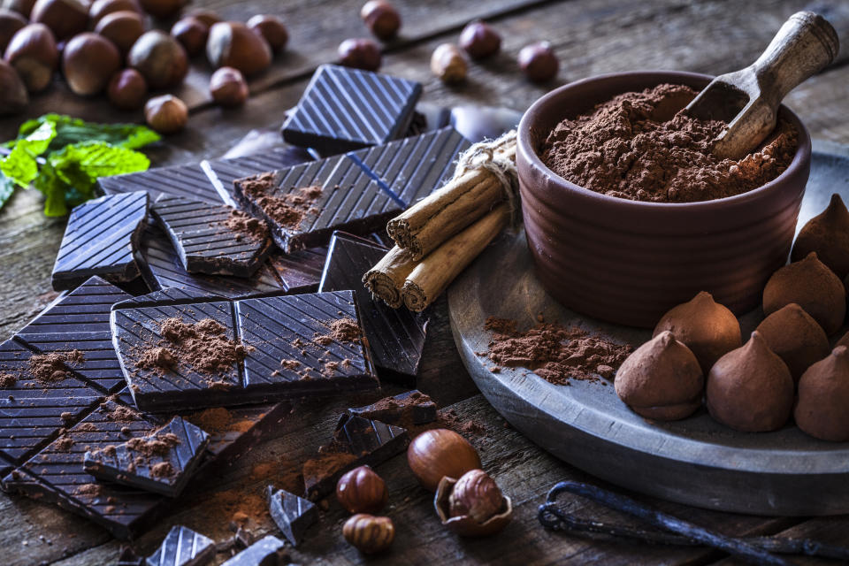 Rustic wooden table filled with ingredient for preparing homemade chocolate truffles. The composition includes dark chocolate truffles, chocolate bars, cocoa powder, cinnamon sticks, vanilla beans, mint leaves and hazelnuts. Predominant color is brown.