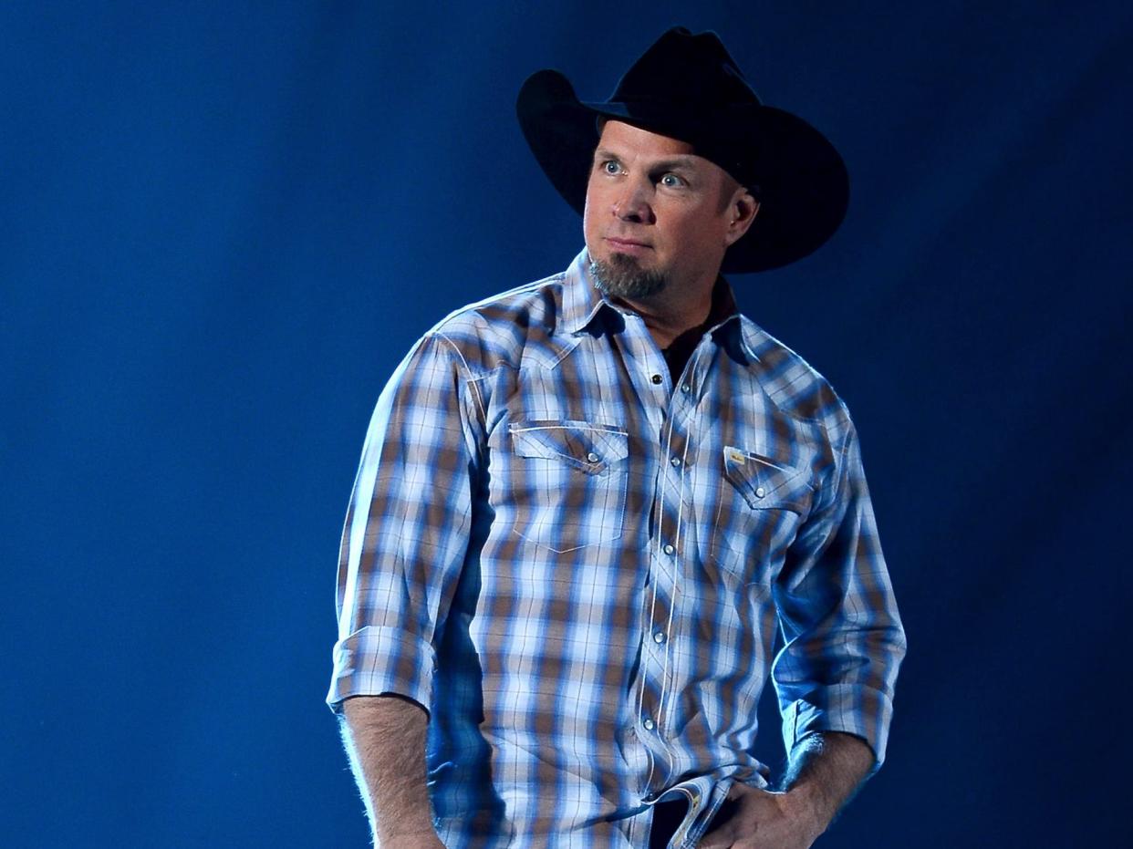 Garth Brooks standing on stage with dark blue lighting while wearing a blue and grey plaid shirt and a black cowboy hat.